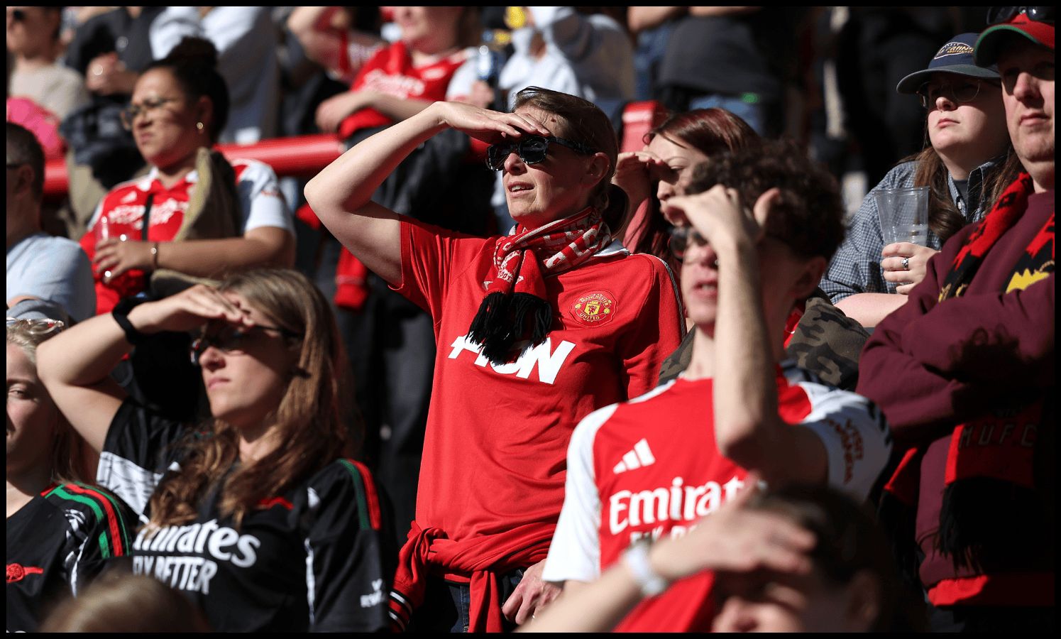 Man United and Arsenal women fans shield their eyes from the sun while watching a game from the stands.