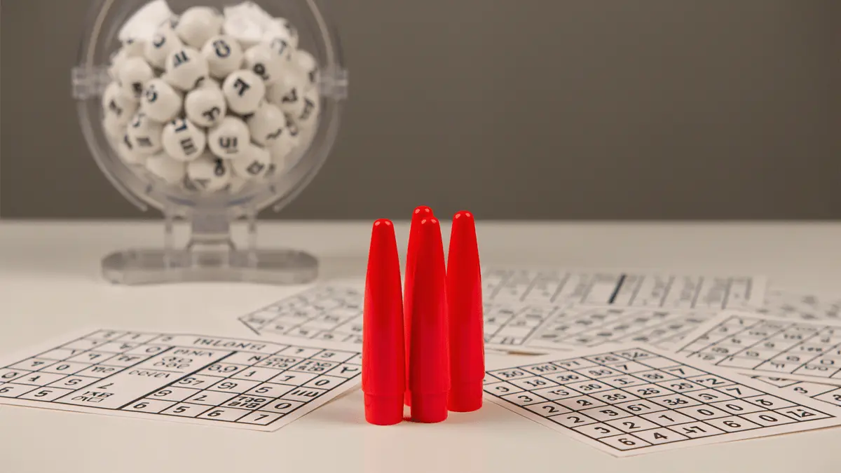 Red bingo dabbers and traditional bingo cards on white table with numbered ball cage in background