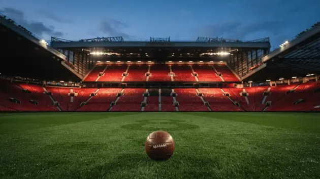 Old Trafford stadium interior with red seats under floodlights and football on pitch