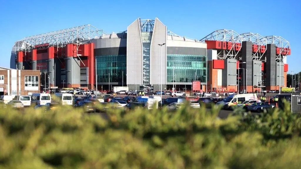 Exterior view of Old Trafford stadium with clear sky and parking area in front.
