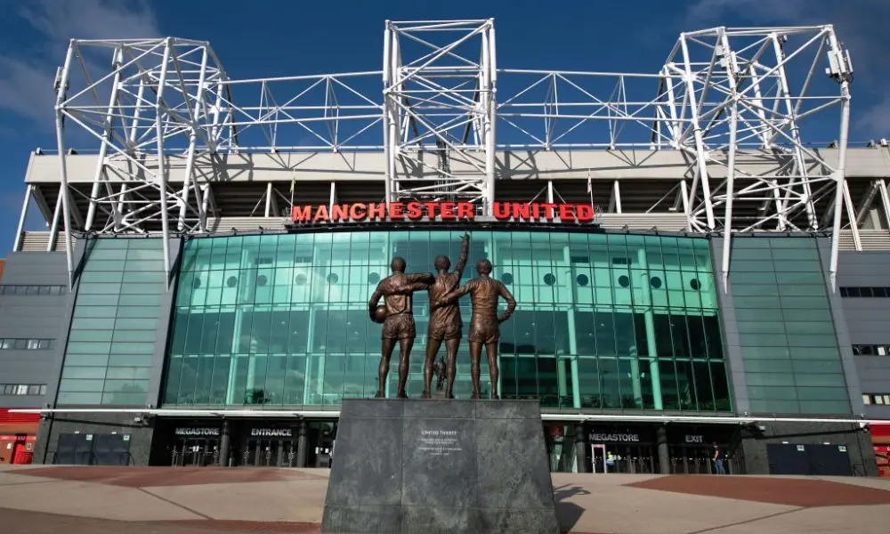Exterior view of Old Trafford stadium with statues of players in front.