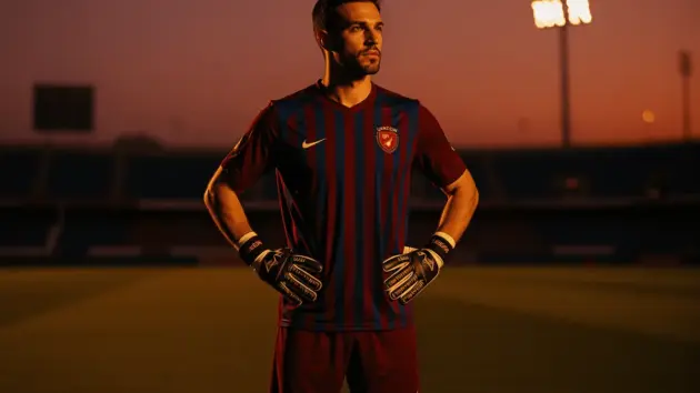 Goalkeeper in maroon jersey stands alone on Turkish stadium pitch at dusk in contemplative pose