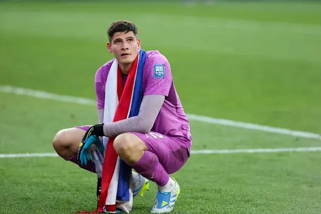 Goalkeeper in pink kit kneeling with a tricolor scarf on the field.
