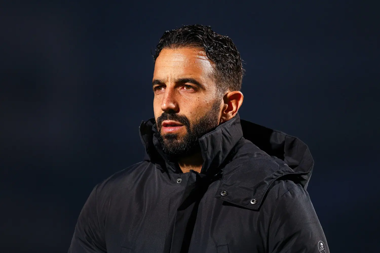 Ruben Amorim, wearing a black coat, looks thoughtfully off-camera during a match.
