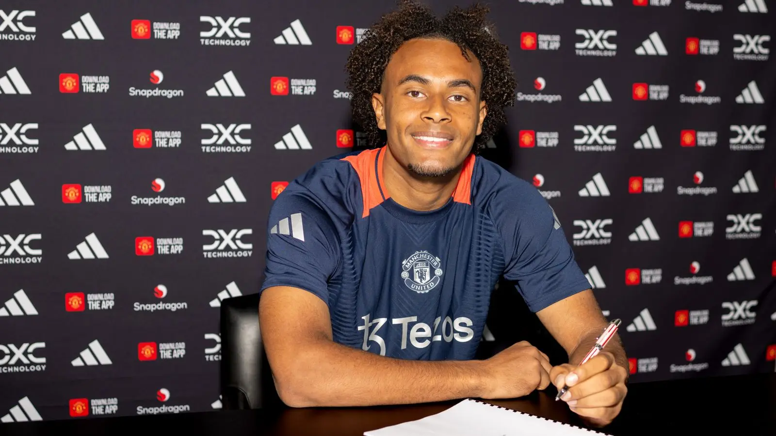 Joshua Zirkzee smiling and holding a pen while signing a contract at Manchester United