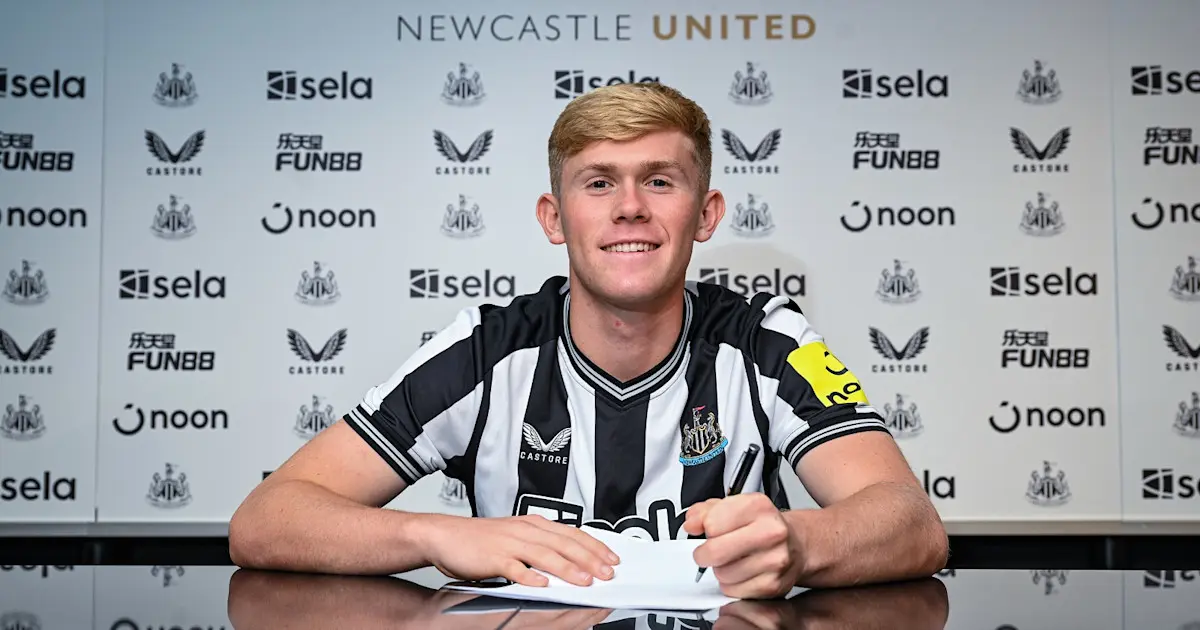 Lewis Hall smiling while signing a contract in a Newcastle United jersey against a club sponsor backdrop.