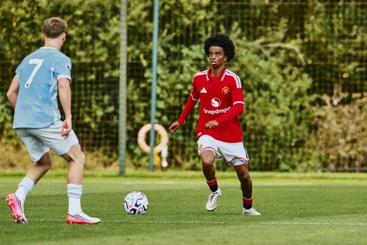 Manchester United player in red kit facing a Manchester City player in light blue kit during a youth match.