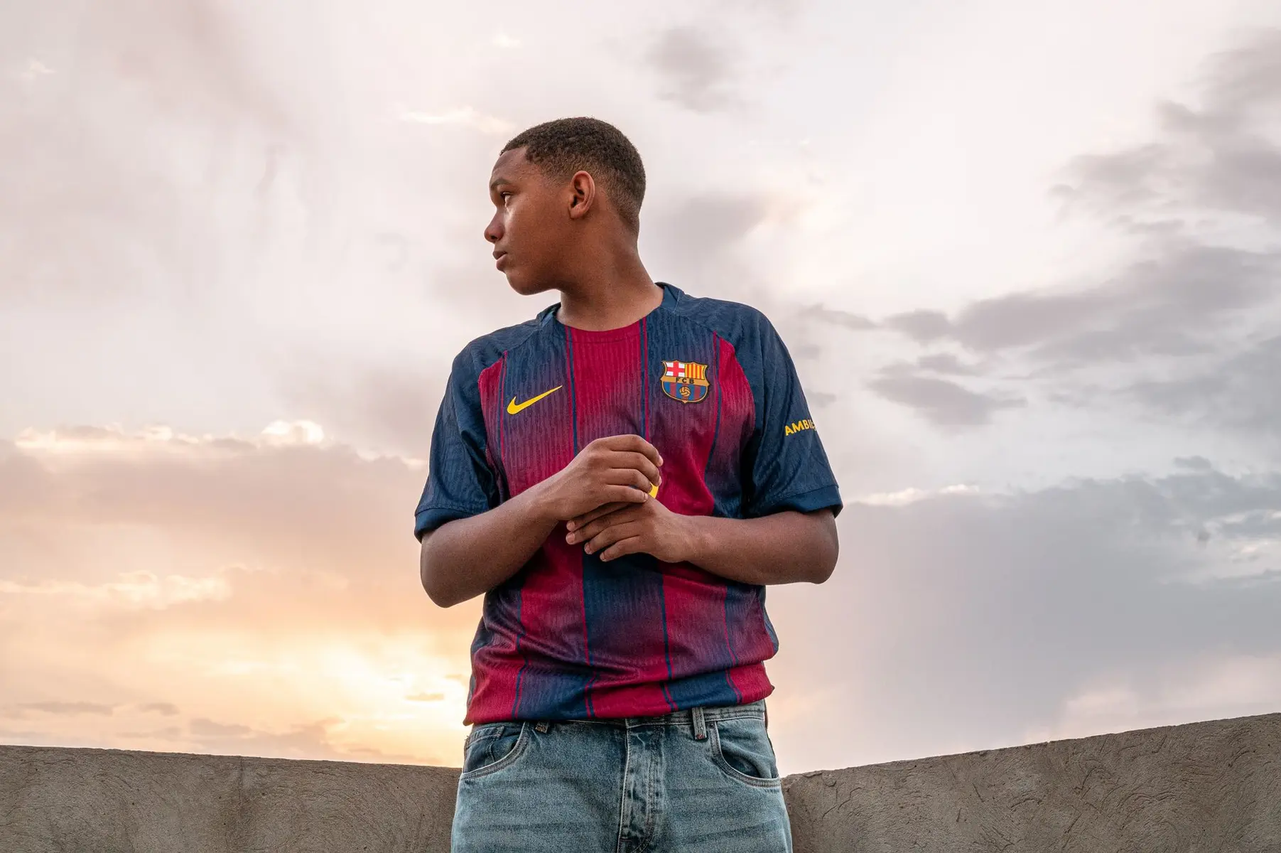 A young man wearing a vintage FC Barcelona soccer jersey standing against a cloudy sunset sky.