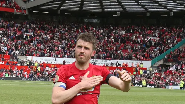 Michael Carrick in a red Manchester United kit standing on a football pitch with a crowd in the background.