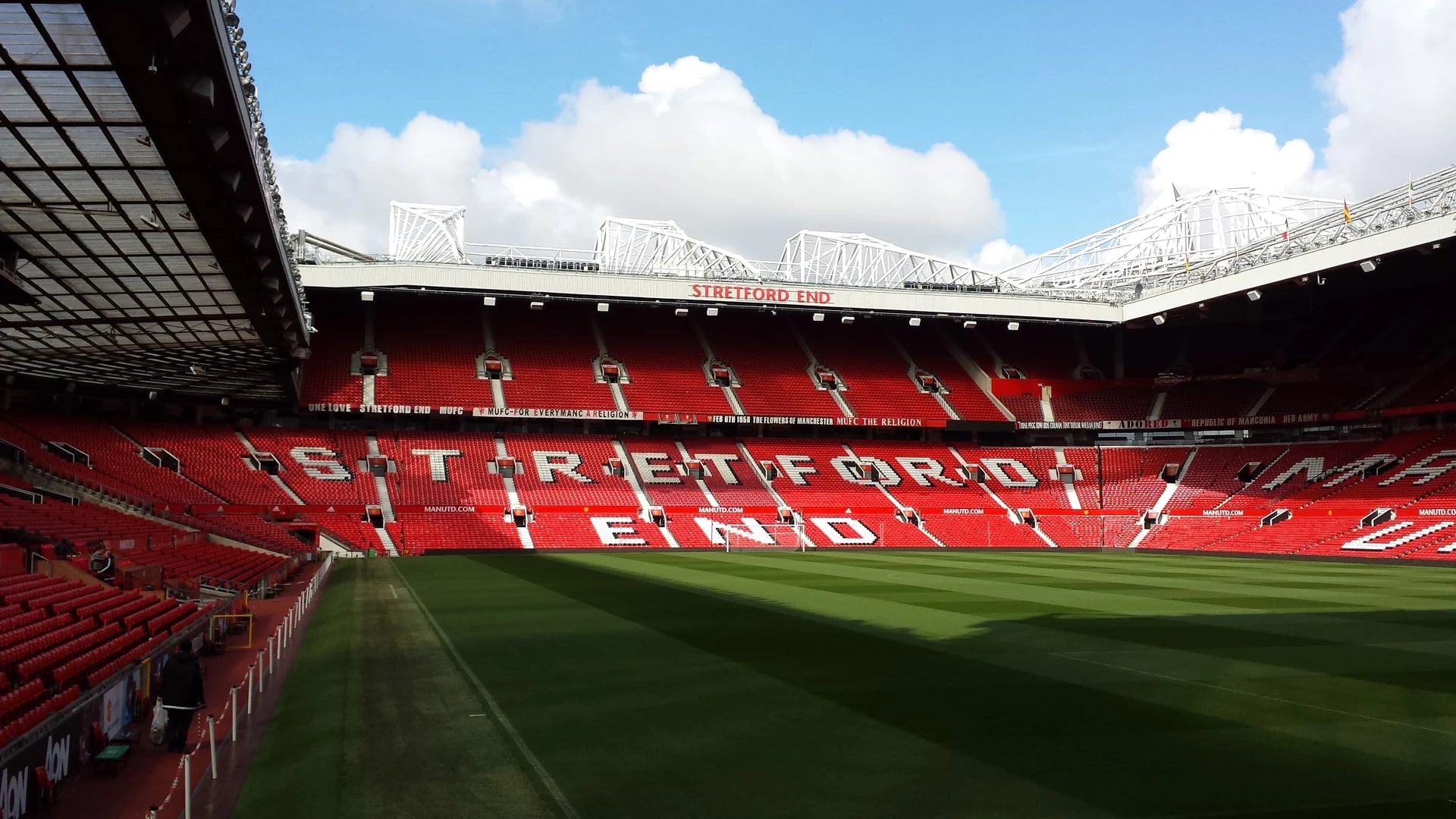A wide view of the empty red seats and green pitch at Old Trafford stadium, focusing on the Stretford End stand.
