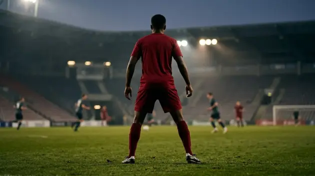 Young centre-back in a red shirt controlling the ball under floodlights during a football match