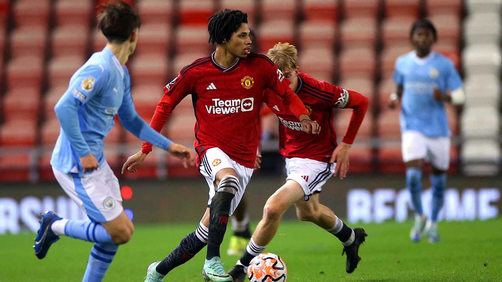 Manchester United Under-21 players in red kits competing for the ball against opponents in light blue kits.