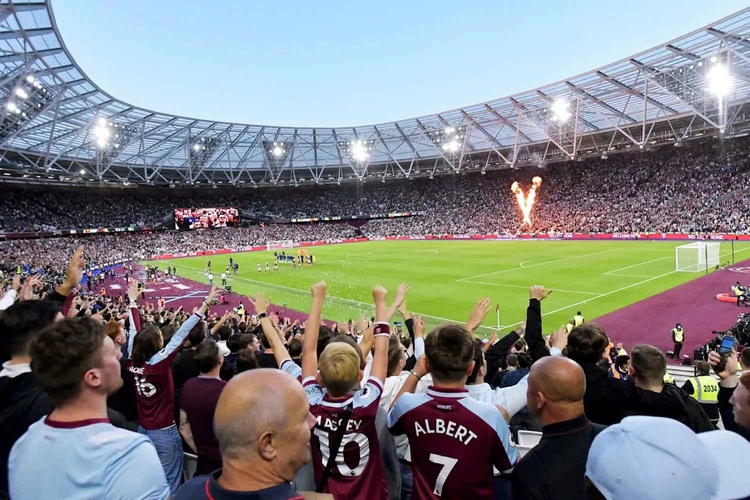 Fans cheering in the stands at London Stadium during a West Ham United match with pyrotechnics on the pitch.