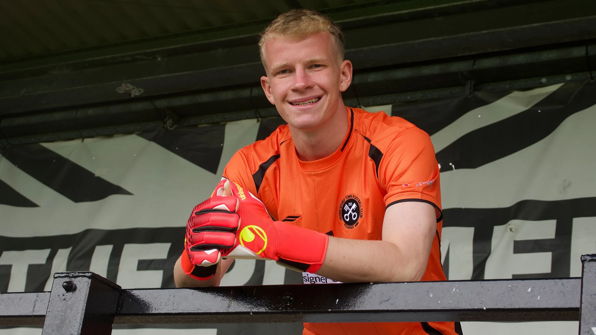 Will Murdock wearing an orange goalkeeper jersey and gloves, leaning on a black railing