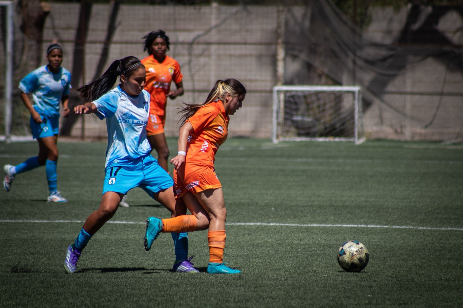 Two women's football players in sky blue and orange kits challenge for the ball during a match