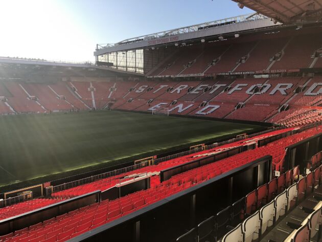 Empty red seats of Old Trafford's Stretford End overlooking the pitch in sunlight