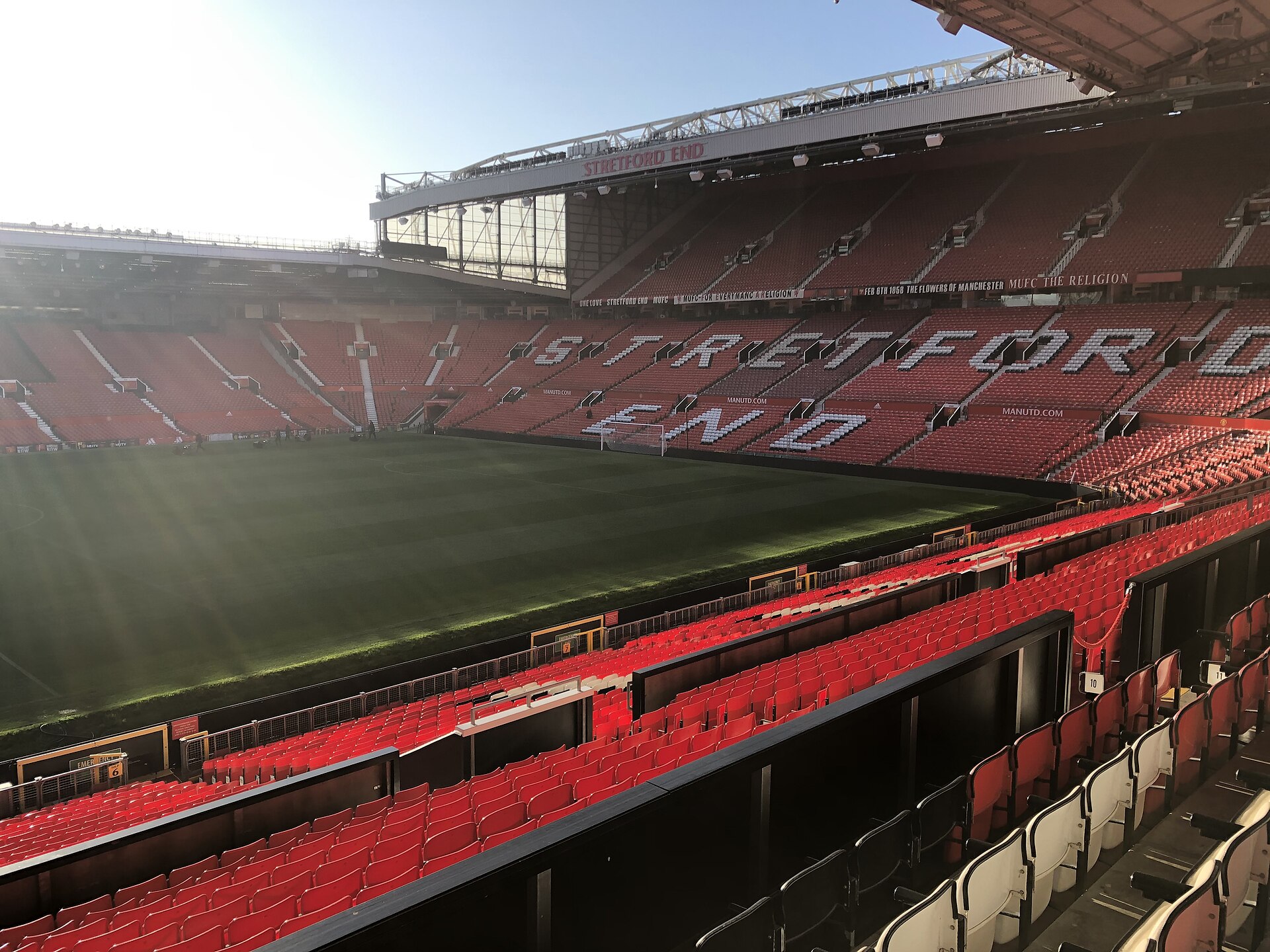 Empty red seats of Old Trafford's Stretford End overlooking the pitch in sunlight