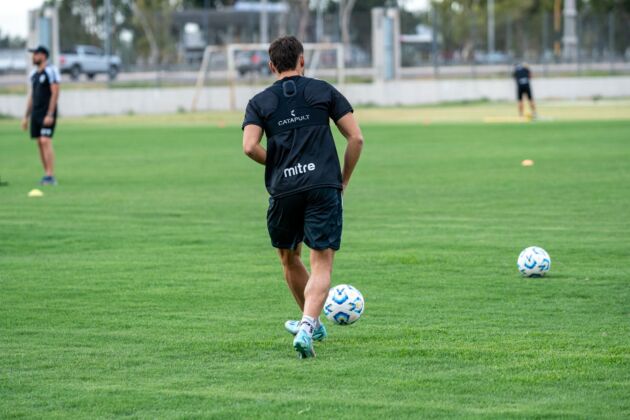 A footballer in black training kit runs with the ball across an empty training pitch